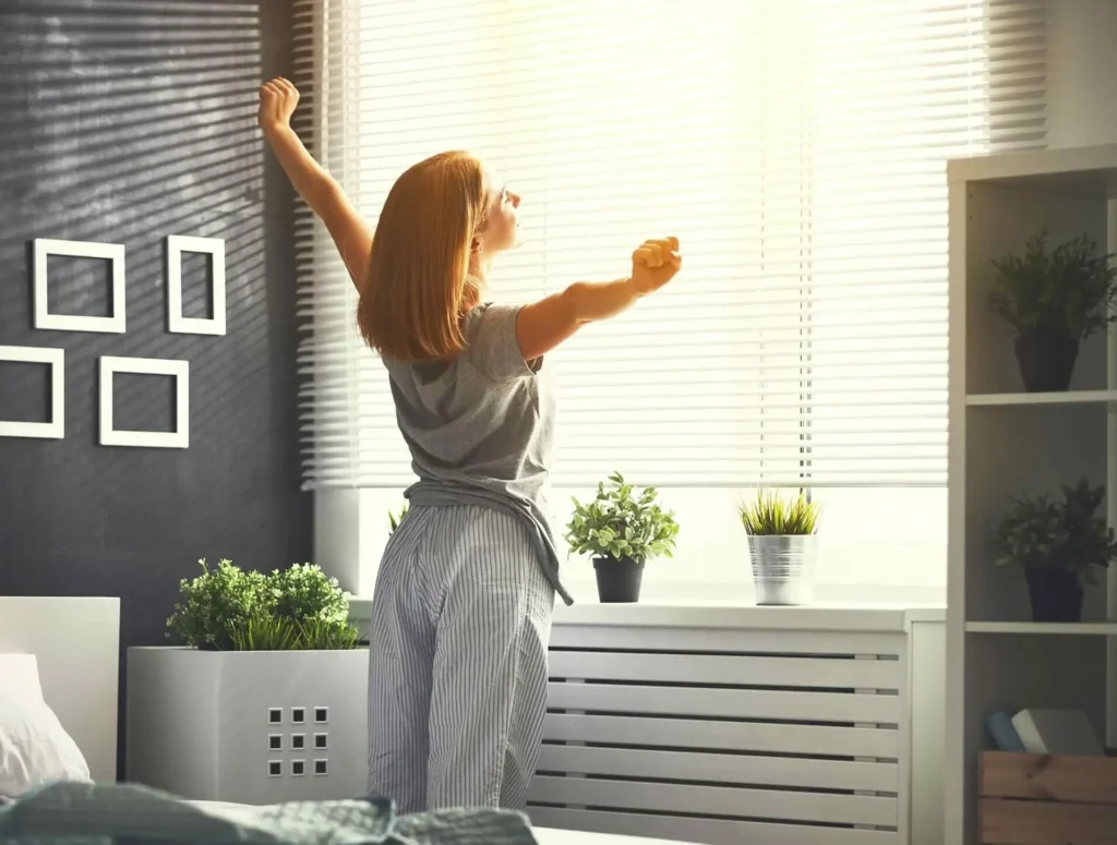 Dame står bak innsynsdempende solskjerming i pysj og føler varmen fra solen